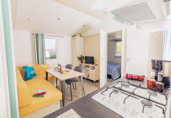Mobil-home lumineux Comfort BAHIA avec cuisine, coin repas, canapé jaune et vue sur la chambre.