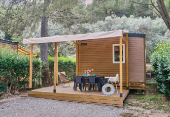 Mobile Home Premium mit Holzterrasse, Tisch und Stühlen im Freien, umgeben von grünen Büschen und Bäumen.