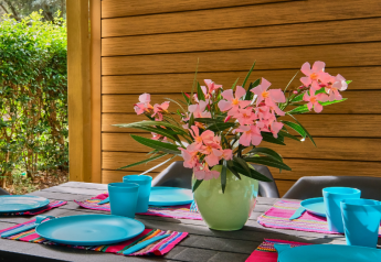 Gedeckter Gartentisch mit blauen Tellern, Bechern und Vase mit rosa Blumen am Mobilheim La Vallée Heureuse.