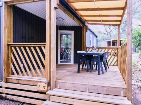 Covered deck of Mobile Home Premium, featuring wood railing, table, four chairs, and wooded view.