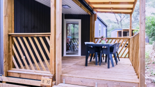 Covered deck of Mobile Home Premium, featuring wood railing, table, four chairs, and wooded view.