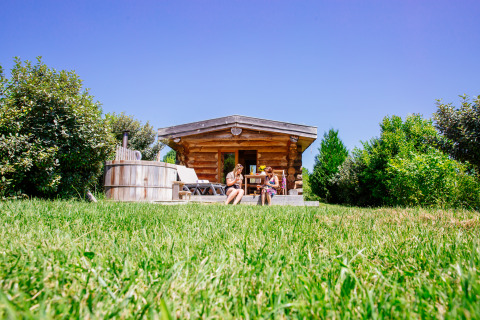 Due persone si rilassano davanti al Log cabin «Trappeur» con tinozza da bagno al Domaine EcÔtelia, Francia, immersi nel verde.