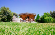 Two people relax outside the Log cabin ‘Trappeur’ with hot tub at Domaine EcÔtelia, France, surrounded by greenery.