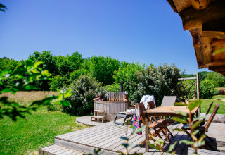 Holzterrasse mit Esstisch und Badezuber vor einem üppigen Wald bei Log cabin «Trappeur» im Domaine EcÔtelia, Frankreich.