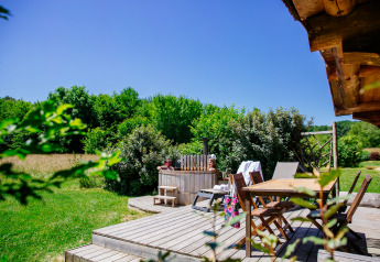 Wooden deck with dining table and hot tub next to lush greenery at Log cabin «Trappeur» in Domaine EcÔtelia, France.