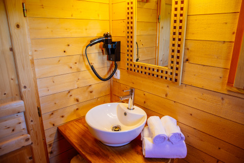 Salle de bain en cabane avec vasque ronde, miroir, sèche-cheveux mural et serviettes roulées sur plan en bois.