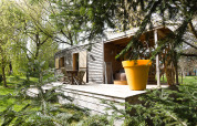 Cabaña de madera con terraza, mobiliario de jardín y una gran maceta naranja rodeada de árboles verdes.