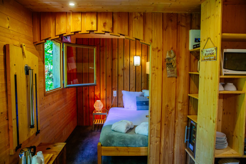 Cozy lodge interior with a double bed, wood paneling, open window, and a kitchenette with microwave.