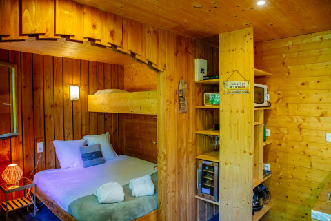 Wood-paneled lodge room featuring a double bed, bunk bed, shelves, minibar, and cozy lighting.
