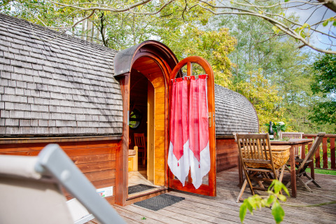 Caravana de madera con puerta abierta y cortina roja en terraza, comedor exterior en Domaine EcÔtelia, Francia.