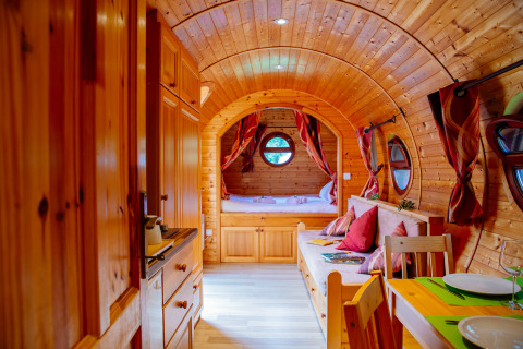 Interior de una acogedora cabaña de madera con techo curvo, mesa de comedor, sofá y cama al fondo.