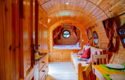 Interior de una acogedora cabaña de madera con techo curvo, mesa de comedor, sofá y cama al fondo.