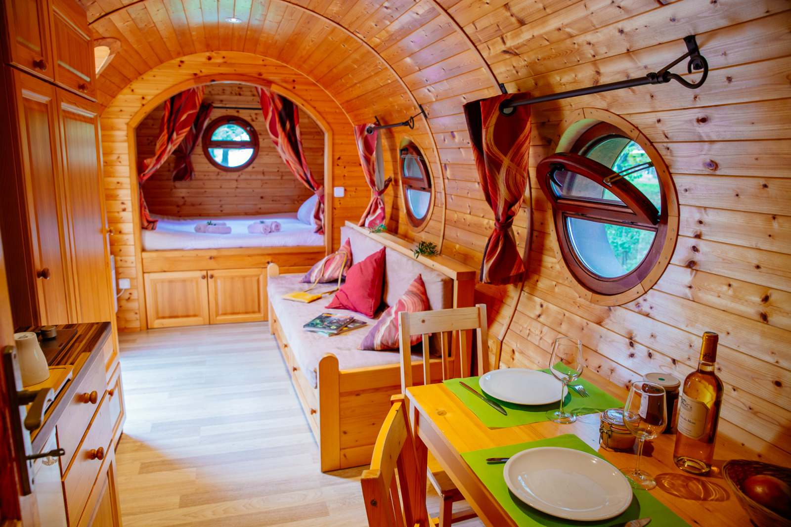 Gezellige houten hut met ronde ramen, eettafel, bank en bed, allemaal warm en sfeervol ingericht.
