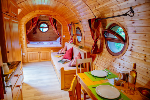 Gemütliche Holzhütte mit runden Fenstern, Esstisch, Sofa und Bett im Hintergrund, warm dekoriert.