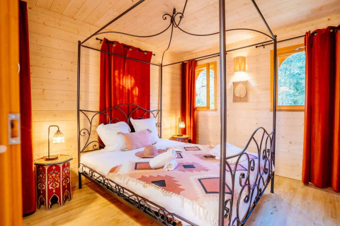 Zimmer in der Lodge 'Riad' im Domaine EcÔtelia in Frankreich mit Himmelbett, roten Vorhängen und Holzdekor.