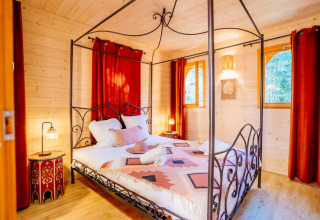 Zimmer in der Lodge 'Riad' im Domaine EcÔtelia in Frankreich mit Himmelbett, roten Vorhängen und Holzdekor.