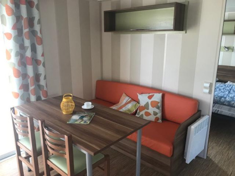 Essbereich im Mobilheim Comfort PMR HELIOS mit Holztisch, orangem Sofa und bedrucktem Vorhang in Frankreich.