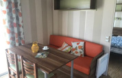 Essbereich im Mobilheim Comfort PMR HELIOS mit Holztisch, orangem Sofa und bedrucktem Vorhang in Frankreich.