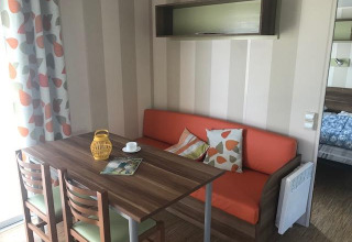 Zona pranzo nella casa mobile Comfort PMR HELIOS con tavolo in legno, divano arancione e tende fantasia in Francia.