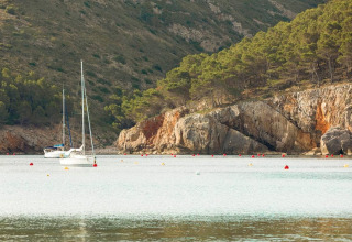 Two sailboats are anchored in calm water surrounded by rocky hills and pine trees near L'Escala, Spain.