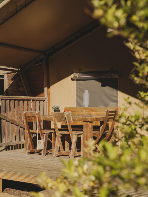 Houten eettafel met stoelen op veranda voor safaritent bij Lodge Family Plus Montgó, omringd door groen.