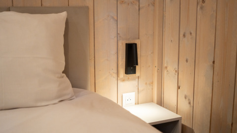 Nahaufnahme von Bett, Kissen, Holzwand und Nachttisch in der Ecolodge - Care accessible im Hameau de l'Ourthe, Belgien.