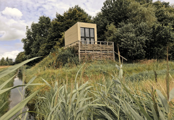 Tiny house est installée sur une plateforme surélevée, entourée d’herbe et d’arbres près d’un canal.