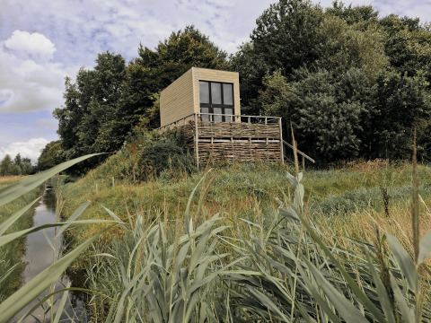 Et lille hus kaldet Tiny house ligger på en hævet platform omgivet af græs og træer ved en kanal.