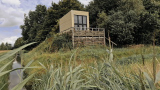 Et lille hus kaldet Tiny house ligger på en hævet platform omgivet af græs og træer ved en kanal.
