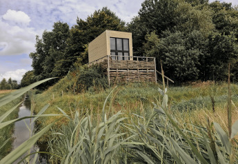 Tiny house est installée sur une plateforme surélevée, entourée d’herbe et d’arbres près d’un canal.
