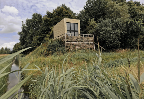 Tiny house staat verhoogd, omringd door gras en bomen, aan de oever van een kleine waterloop.