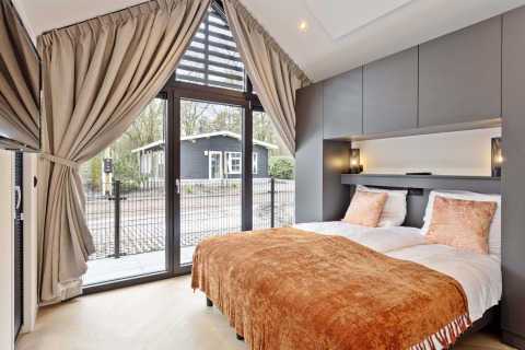 Dormitorio moderno en lodge con grandes ventanas, cortinas, cama doble y vistas al jardín y casa exterior.