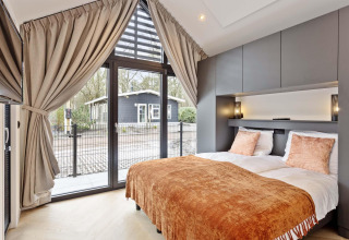 Dormitorio moderno en lodge con grandes ventanas, cortinas, cama doble y vistas al jardín y casa exterior.