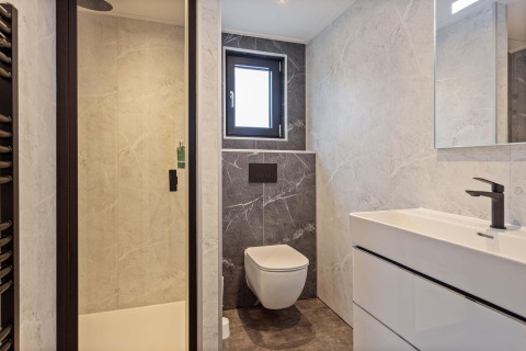 Baño moderno en una cabaña con inodoro suspendido, ducha de vidrio, ventana pequeña y lavabo blanco.