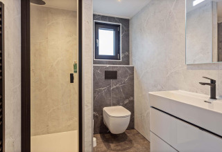 Baño moderno en una cabaña con inodoro suspendido, ducha de vidrio, ventana pequeña y lavabo blanco.