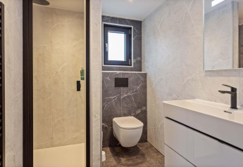 Baño moderno en una cabaña con inodoro suspendido, ducha de vidrio, ventana pequeña y lavabo blanco.