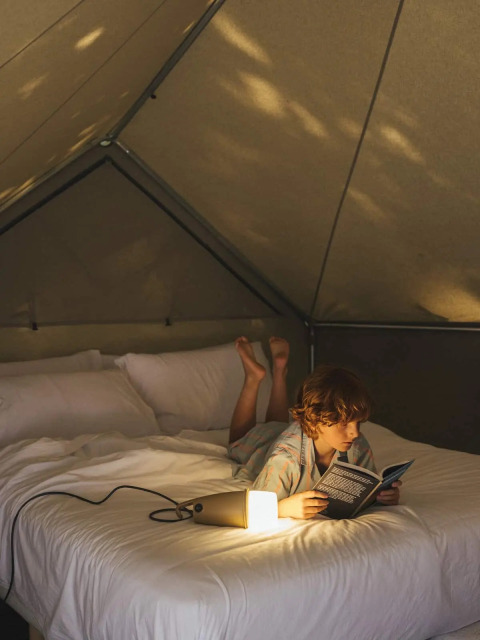 Un enfant lit un livre allongé sur un lit dans une tente safari, éclairée par une petite lampe conviviale.