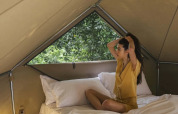 Mujer sentada en una cama dentro de una lujosa tienda safari con ventana triangular y sábanas blancas.