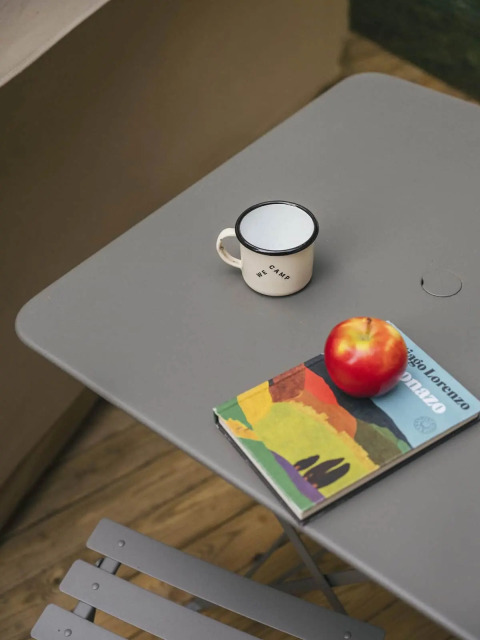 Table grise sous tente safari avec une tasse émaillée, une pomme rouge et un livre coloré dessus.