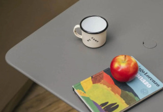 Table grise sous tente safari avec une tasse émaillée, une pomme rouge et un livre coloré dessus.