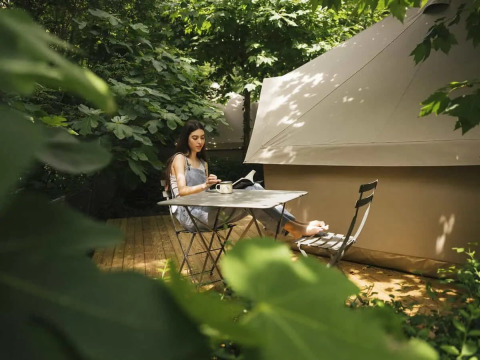 Femme assise à une table près d'une tente safari, entourée de verdure et de lumière naturelle.