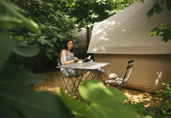 Femme assise à une table près d'une tente safari, entourée de verdure et de lumière naturelle.