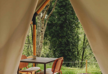 Vista da una tenda safari glamping con ingresso aperto, tavolo e sedie, circondata da alberi verdi in Spagna.