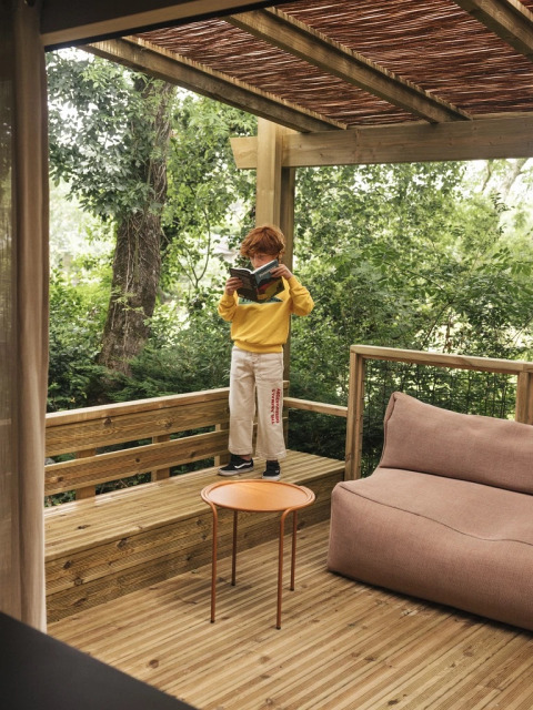 Kind leest een boek op het houten terras van Mobil Home Family Plus bij Wecamp San Sebastián, te midden van groen.