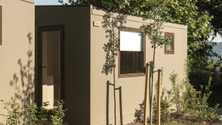 Lodge Mobile Home Couple Plus à façade beige, entouré de verdure et d’arbres, avec un sentier devant.