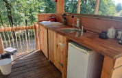 Outdoor wooden kitchenette with sink, mini fridge, and forest view at Bell Tent lodge, Campsite Junora, France.
