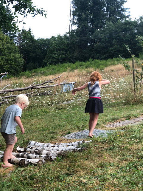 Two children walk barefoot on a log path at a glamping site, surrounded by green grass and trees.