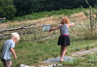 Two children walk barefoot on a log path at a glamping site, surrounded by green grass and trees.