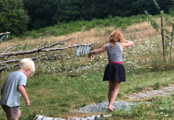 Dos niños caminan descalzos sobre un sendero de troncos en un alojamiento glamping rodeado de naturaleza.