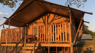 Sunlit luxury and spacious eco-lodge safari tent with wooden deck and railing set in natural surroundings.